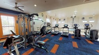 This single-floor fitness facility at Spring Garden Drive in Fort Worth, Texas, offers a comprehensive workout environment designed to meet a wide range of athletic and fitness needs. The space is divided into two interconnected areas: a fully equipped gym and an adjacent indoor basketball court.

The fitness area features bold orange and navy blue walls accented by a vibrant blue-and-orange patterned carpet, creating an energetic and motivating atmosphere. Ceiling fans provide airflow throughout the space, which is illuminated by recessed lighting. Equipment includes a multi-function cable machine, a full rack of dumbbells, kettlebells in varying weights, spin bikes, treadmills, stair climbers, and elliptical machines — catering to both cardio and strength training routines. A mat station and sanitation dispenser are also available for convenience.

Adjacent to the gym, the indoor basketball court boasts soaring ceilings and abundant natural light streaming through a row of high windows and an arched transom window. NBA team logos adorn the bright white walls, adding a spirited, sport-inspired character. A scoreboard and basketball hoops complete the court. A metal railing separates the fitness zone from the court below, and track lighting overhead ensures excellent visibility. Together, these spaces provide a dynamic and well-rounded athletic facility.