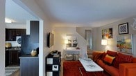 This three-floor residence offers a thoughtfully arranged living environment with a design that balances functionality and style. The main living area serves as the heart of the home, featuring neutral wall tones complemented by warm accent colors. A bold red area rug anchors the seating arrangement, where a vibrant orange sofa adds a striking focal point against the understated backdrop. A wall-mounted flat-screen television is positioned above a sleek black storage chest, providing both entertainment and organizational utility.

A white console table styled with decorative antler accents and table lamps introduces an eclectic, contemporary aesthetic to the space. Woven storage baskets beneath the console add a practical yet textured element to the décor. The open layout allows for a natural sightline through the space, creating a sense of depth and connectivity between areas.

A staircase visible in the background provides access to the upper levels, reinforcing the multi-floor nature of the home. Soft, layered lighting from multiple lamp sources gives the living area a warm and inviting ambiance. Overall, the space presents a well-curated interior with a mix of modern and eclectic design influences.