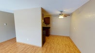 This single-floor space offers a straightforward, functional layout designed for comfortable everyday living. The main living area is finished with warm light hardwood flooring that extends throughout, creating a bright and cohesive feel underfoot. A ceiling fan with an integrated light fixture is centrally positioned overhead, providing both air circulation and ambient lighting to the room. The walls are painted in a neutral tone, keeping the space feeling open and versatile.

Adjacent to the living area, the kitchen is partially separated by a partial wall divider, giving it a defined presence while maintaining a connection to the main space. The kitchen is outfitted with rich, dark wood cabinetry paired with dark countertops, offering a pleasant contrast to the lighter tones of the surrounding walls and flooring. A range or cooktop is visible, indicating a fully functional cooking area. The textured ceiling adds a subtle architectural detail throughout the space.

Overall, this single-floor layout presents a clean, move-in ready environment with practical features, neutral finishes, and a cohesive design that offers a solid foundation for personalization.
