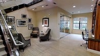 This refined two-story space makes a strong first impression with its elegantly appointed lobby area, designed to balance professionalism with comfort. The main level features a well-furnished lounge seating area with sleek, dark-framed accent chairs and coordinating side tables, arranged to encourage both relaxation and conversation. Framed artwork adorns the warm, neutral-toned walls, adding a curated, gallery-like quality to the space.

Flooring throughout is covered in a neutral, linear-patterned carpet that complements the understated color palette. Recessed lighting illuminates the interior evenly, while a staircase with a bold dark railing leads to the upper floor, adding a striking vertical element to the design.

A standout feature is the full-height glass partition wall that separates an adjacent room, maintaining an open, airy feel while defining distinct functional zones. This glass-enclosed area benefits from generous natural light streaming through large windows that frame views of lush greenery outside, creating a bright and inviting atmosphere.

The overall ambiance is clean, modern, and professional, making this space well-suited for a business or hospitality environment. The thoughtful combination of architectural details, natural light, and contemporary décor gives the space a sophisticated yet approachable character across both its levels.
