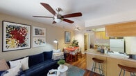 This single-floor residence offers a thoughtfully designed layout that seamlessly connects the main living areas. The open-concept living, dining, and kitchen zone forms the heart of the home, anchored by rich hardwood-style flooring that extends throughout the space. The living area features a ceiling fan with light fixture and is bathed in warm ambient lighting, creating an inviting atmosphere. A breakfast bar with counter seating bridges the kitchen and living space, providing both functionality and a casual dining option. The kitchen is appointed with warm wood cabinetry, a gas range, refrigerator, and a stainless steel sink, with ample upper and lower cabinet storage. A dedicated dining area accommodates a full table and chairs, ideal for everyday meals or entertaining. Both bedrooms are comfortably sized, each featuring ceiling fans, hardwood-style flooring, and closets with hanging storage. One bedroom includes a window with blinds and draping curtains, allowing for natural light control. The bathroom connects directly to one of the bedrooms and includes a closet, a vanity, and a towel bar. Framed artwork is displayed throughout, contributing to a curated and cohesive aesthetic across the entire residence.
