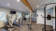 This fitness center offers a comprehensive workout environment designed to accommodate a variety of exercise routines. The space is defined by a distinctive exposed wood beam running across the ceiling, adding a warm architectural contrast to the otherwise clean, light gray walls and white trim throughout. Recessed lighting illuminates the room evenly, complementing the natural light that enters through windows along the exterior wall.

The flooring consists of durable, speckled rubberized material suited for high-traffic use and heavy equipment. A full-length mirrored wall on one side visually expands the space while allowing users to monitor their form. The cardio section is well-stocked with multiple treadmills, elliptical trainers, and a stair climber, while the strength training area includes a cable machine, multi-function weight station, adjustable benches, a power tower, and free weights. A wall-mounted flat-screen television adds convenience for entertainment during workouts.

A restroom is accessible directly from the fitness area, and a pass-through window connects to an adjacent room. The overall layout is efficiently organized, providing ample space to move between stations comfortably.