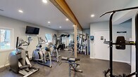 This fitness center offers a comprehensive workout environment designed to accommodate a variety of exercise routines. The space is defined by a distinctive exposed wood beam running across the ceiling, adding a warm architectural contrast to the otherwise clean, light gray walls and white trim throughout. Recessed lighting illuminates the room evenly, complementing the natural light that enters through windows along the exterior wall.

The flooring consists of durable, speckled rubberized material suited for high-traffic use and heavy equipment. A full-length mirrored wall on one side visually expands the space while allowing users to monitor their form. The cardio section is well-stocked with multiple treadmills, elliptical trainers, and a stair climber, while the strength training area includes a cable machine, multi-function weight station, adjustable benches, a power tower, and free weights. A wall-mounted flat-screen television adds convenience for entertainment during workouts.

A restroom is accessible directly from the fitness area, and a pass-through window connects to an adjacent room. The overall layout is efficiently organized, providing ample space to move between stations comfortably.