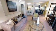 The Lenox Classic is a two-story, 2-bedroom, 2-bathroom residence designed with an open, flowing layout that connects the main living areas seamlessly. The living room serves as a welcoming centerpiece, highlighted by a bold teal accent wall, a fireplace, and a wall-mounted television flanked by shelving units. Sliding glass doors lead to a private balcony, filling the space with natural light and offering an outdoor retreat with seating. Carpeted flooring and patterned area rugs add warmth and texture throughout.

The dining area is positioned adjacent to the living room and features a glass-top table surrounded by upholstered blue chairs. A large mirror on the dining room wall enhances the sense of space and reflects light throughout the area. Full-length patterned drapes frame the sliding doors, adding a polished finishing touch.

The kitchen is accessible from the dining area and includes white cabinetry, a full-size refrigerator, and a breakfast bar with bar stool seating—ideal for casual dining. The bar counter connects the kitchen to the living space, reinforcing the open-concept design. The overall ambiance is contemporary and well-appointed, with cohesive color choices in navy, gold, and white tones carried throughout the home.