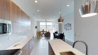 Located at 2304 Hillsborough in Raleigh, NC, this single-floor space offers a thoughtfully designed open-concept layout that seamlessly connects the kitchen, dining, and living areas. The kitchen is outfitted with sleek flat-front cabinetry in a warm wood finish, light quartz countertops with a clean waterfall edge, a subway tile backsplash, and modern stainless steel fixtures. A stylish copper pendant light anchors the kitchen space, adding a warm contemporary accent. The dining area accommodates a full table and chairs, positioned conveniently between the kitchen and living room for effortless entertaining. The living room is bright and inviting, furnished with comfortable seating and bathed in natural light from expansive floor-to-ceiling windows that frame views of the surrounding urban landscape. A ceiling fan overhead and recessed lighting throughout ensure year-round comfort and a well-lit interior. The flooring is consistent large-format tile throughout, contributing to the clean, modern aesthetic. The white walls and high ceilings enhance the sense of openness and airiness across the entire space. Overall, the unit presents a polished, contemporary design well-suited for urban living in the heart of Raleigh.
