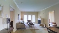 This single-floor space centers around a generously sized living room that combines comfort and classic design elements. The room is finished with neutral beige walls and soft carpeting, contributing to a warm, understated aesthetic throughout. Crown molding lines the ceiling, adding a refined architectural detail that elevates the overall feel of the space.

A white-mantled fireplace serves as a prominent focal point along one wall, providing both a functional heating element and a decorative centerpiece. Opposite the fireplace, a set of white French doors floods the room with natural light, offering a bright outlook and easy access to what appears to be an outdoor area. The doors enhance the sense of openness, making the living area feel airy and well-connected to its surroundings.

The layout comfortably accommodates a full seating arrangement, including a sofa, accent chair, and ottoman, all centered around a rectangular coffee table atop a layered area rug. A table lamp and decorative wall-mounted artwork add finishing touches. The space is practical, well-lit, and thoughtfully arranged for everyday living.
