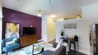 This studio is thoughtfully designed to make efficient use of its open floor plan. The living area serves as the centerpiece of the space, featuring a striking deep purple accent wall that adds a bold visual contrast against the soft cream-toned surrounding walls and white ceiling. A ceiling fan with a light fixture keeps the area comfortable and well-lit. Dark hardwood-style flooring runs throughout, grounding the space with a warm, modern aesthetic.

The living area accommodates a plush sectional sofa, a glass-top coffee table set atop a geometric-patterned area rug, and a wooden media console supporting a flat-screen television. Adjacent to the living area, a narrow bar-style counter with industrial metal stools provides a dedicated dining or workspace, complemented by decorative wall-mounted shelving featuring minimalist branch artwork above.

A hallway leads toward what appears to be a private bathroom and closet area, with white paneled doors providing clean definition between spaces. The layout is open and well-organized, offering a practical and stylish living environment suited for single occupancy.