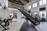 Located at 659 Treeland Dr in Ladson, South Carolina, this two-story fitness center is a well-equipped and thoughtfully designed facility that caters to a comprehensive range of exercise needs. The ground floor houses multiple workout zones, each finished with light wood-style flooring and neutral walls that create a clean, energizing atmosphere. Large windows fitted with white plantation shutters allow ample natural light to flow throughout, with views of the surrounding community, including an outdoor pool area visible from certain vantage points. French doors with glass panels provide additional access points and further brighten the space. Ceiling fans are installed throughout to maintain air circulation, and recessed lighting supplements natural light during evening hours.

The upper level features a dramatic vaulted ceiling adorned with exposed rustic wood beams accented by industrial hardware, adding a bold architectural character. A large motivational mural spanning the full height of the walls reinforces the wellness-focused design intent. A sleek black metal staircase with cable railings connects the two floors, and upper-level windows offer elevated views of the neighborhood. Equipment across both floors includes cable machines, a multi-station weight system, rowing machines, spin bikes, dumbbells, stability balls, and battle ropes, making this a fully functional, multi-use fitness destination.