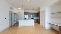This well-appointed single-floor residence showcases a clean, modern aesthetic throughout its open-concept layout. The kitchen serves as the centerpiece of the main living area, featuring dark wood cabinetry that contrasts beautifully with the bright white kitchen island topped with a light stone-style countertop. A sleek pendant light hangs above the island, complemented by a track lighting system that ensures the space is well-lit at all times. Stainless steel appliances — including a French door refrigerator, built-in microwave, and electric range — add a polished, professional feel to the kitchen. A glass tile backsplash in a subtle grey tone ties the design together with refined detail. Light wood-tone flooring runs throughout the main area, contributing to the bright, airy ambiance of the space. Just off the kitchen, a dedicated in-unit laundry area houses a full-size washer and dryer, offering everyday convenience. The layout flows naturally, with additional rooms accessible via clean white interior doors. High ceilings and a predominantly white palette amplify the sense of space and openness, making this a highly functional and visually inviting residence.