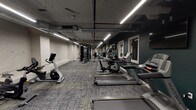 This fitness room offers a comprehensive workout environment designed to accommodate a wide range of exercise needs. The space is organized into distinct zones, allowing users to move seamlessly between cardio, strength, and functional training areas. Along one side of the room, a row of cardio machines — including Matrix treadmills, upright bikes, and a recumbent bike — provides ample options for aerobic exercise. A dedicated strength training area features a multi-station cable machine, while a full dumbbell rack with hex weights and a wall-mounted flat-screen TV anchors another section of the room. A separate open area houses stability balls, medicine balls, and resistance accessories, ideal for stretching and functional fitness routines. The room's industrial aesthetic is defined by exposed ductwork and piping along the ceiling, white-painted brick walls, and patterned low-pile carpet flooring. Linear LED lighting runs throughout, ensuring the space is evenly and brightly lit. Deep teal accent walls add a modern contrast to the white brick, giving the fitness room a polished, contemporary feel. Mirrored panels are incorporated into the design, enhancing the sense of openness. The layout is spacious and thoughtfully arranged to allow for comfortable movement between equipment zones.