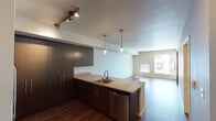 This well-appointed single-floor unit offers a thoughtfully designed open-concept layout that seamlessly connects the kitchen and living area, creating a spacious and functional environment. The kitchen is a standout feature, showcasing rich dark-stained cabinetry with ample storage, granite countertops, and a full suite of stainless steel appliances including a refrigerator, electric range, and built-in microwave. A peninsula with an integrated sink and additional seating space serves as the natural dividing point between the kitchen and the expansive living area. Track lighting and stylish pendant lights illuminate the kitchen, adding a modern touch. The living area is generously sized, bathed in natural light from multiple windows, and finished with warm, rich hardwood flooring that extends throughout the main living spaces. Neutral, light-toned walls complement the overall contemporary aesthetic. The bedroom is comfortably sized, finished with plush carpet, and features a closet with built-in shelving and hanging space. A bathroom with matching dark cabinetry and a sink is also present. The unit's clean lines, modern finishes, and open layout come together to create a bright, inviting, and highly functional living space.