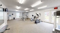 Located at 4211 W Potomac Dr in Greenfield, Indiana, this dedicated fitness center offers a well-organized and comprehensively equipped exercise room designed to support a full range of workout routines. The space is finished with neutral-toned, low-pile carpeting throughout and bright white walls that contribute to a clean, open atmosphere. Overhead, multiple fluorescent panel fixtures provide ample, evenly distributed lighting across the entire room.

Large wall-mounted mirrors are positioned strategically throughout the space, enhancing the sense of depth and allowing users to monitor their form during exercise. The cardio section features treadmills, an upright stationary bike, a recumbent bike, and an elliptical trainer, offering a variety of aerobic options. A multi-station Hoist strength training machine serves as the centerpiece for resistance and weight training, complemented by a set of kettlebells, a dumbbell rack, and resistance bands with suspension trainers mounted on the wall.

Additional accessories include rolled exercise mats and a weight bench for stretching or supplemental exercises. Windows allow for natural light to enter the room, and a wall-mounted television adds an entertainment option for users. The overall layout is thoughtfully arranged to maximize usable floor space while keeping equipment accessible and the environment motivating.