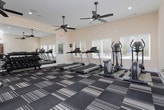 The fitness center at 1000 Hidden Chase in Stone Mountain, Georgia, is a well-equipped, single-floor facility designed to support a comprehensive workout routine. The space is finished with a stylish patterned carpet in neutral tones of black, gray, and beige, providing both comfort and a modern aesthetic throughout.

The cardio section is outfitted with multiple treadmills and elliptical machines, arranged to allow ample space between equipment. A dedicated strength training area features a multi-station cable machine with an adjustable bench, accommodating a wide range of resistance exercises. A full rack of dumbbells in graduated weights adds further versatility for free weight training.

A floor-to-ceiling mirror runs along one wall, visually expanding the space and allowing users to monitor their form during workouts. Multiple windows fitted with blinds allow natural light to flow in from multiple sides, offering views of the surrounding outdoor area. Several ceiling fans are mounted throughout, ensuring consistent airflow and a comfortable environment during exercise.

Recessed lighting supplements the natural light, keeping the space well-lit at all times. The facility is accessible via a dedicated exterior door, offering convenient entry and exit. Clean, neutral-toned walls and white trim give the space a bright, welcoming appearance that is both functional and inviting.