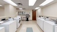 This shared laundry facility is designed for functionality and ease of use, offering a well-organized layout across a single floor. The space is brightened by recessed fluorescent panel lighting set within a drop-tile ceiling, creating a clean, evenly lit environment throughout.

The main laundry area is equipped with multiple front-loading and top-loading washing machines, alongside a bank of stacked dryers lining one wall. The stacked dryer units maximize vertical space, providing ample drying capacity. Each machine features coin-operated controls, and a dedicated coin-change kiosk is conveniently positioned within the room, making transactions straightforward for users.

Wire laundry carts on wheels are available throughout the space for transporting loads, and a chair is provided for those waiting. The floors feature light-toned vinyl tile that is easy to maintain, while the cream-colored walls contribute to the facility's clean, utilitarian aesthetic.

A glass-paneled interior window along one wall allows for visibility into an adjacent room, adding openness to the corridor-style entry area. A fire extinguisher and safety signage are clearly mounted, reflecting attention to safety standards. Overall, the space is practical, well-maintained, and efficiently designed to serve a shared-use community.
