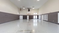 This single-floor indoor sports facility is designed to accommodate a range of athletic and recreational activities, centered around a full-size basketball court. The space is defined by its impressively high ceilings, which allow for unrestricted play and contribute to an open, airy atmosphere throughout the interior.

The court features a polished concrete floor that reflects the overhead lighting, enhancing visibility across the entire playing surface. A regulation basketball backboard and hoop are mounted on the far wall, making the space well-suited for organized games and casual play alike.

The walls are finished in a two-tone treatment, with a gray lower section and a lighter upper portion, creating a clean and functional aesthetic. Natural light enters through a series of windows positioned high along the walls, supplemented by industrial-style ceiling-mounted light fixtures that ensure consistent illumination.

Along the perimeter, multiple doorways provide convenient access points, and additional wall-mounted equipment, including a speaker system and climate control unit, reflects the facility's attention to functionality. The overall design prioritizes practicality and performance, making it a versatile space for sporting and community use.
