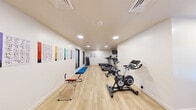 This fitness center offers a clean, modern environment designed to support a well-rounded workout routine. The space is laid out across a single level, with light wood-style flooring running throughout and crisp white walls that contribute to an open, airy atmosphere. Recessed lighting provides consistent, bright illumination across the entire area.

The cardio section is equipped with multiple treadmills and stationary cycling bikes, positioned along the walls to maximize usable floor space. A wall-mounted television is positioned for viewing during cardio sessions. Adjacent to the cardio equipment, a tiered dumbbell rack holds a comprehensive range of free weights, catering to strength training needs. Exercise posters and instructional charts are mounted on the walls, offering guidance for various workouts.

The open floor area accommodates a collection of stability balls in multiple sizes, a yoga or exercise mat, and additional functional fitness accessories. A water fountain and waste receptacle are conveniently located near the entrance, which features a set of glass-panel double doors providing a welcoming entry point. Windows allow natural light to supplement the overhead lighting, enhancing the overall brightness of the space. The layout is efficient and uncluttered, creating a practical and inviting environment for exercise.