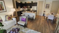 This well-appointed single-floor space showcases an open-concept layout that seamlessly connects the living and kitchen areas, creating a cohesive and functional flow. Rich hardwood flooring runs throughout, anchoring the space with warmth and elegance.

The kitchen is a standout feature, outfitted with dark espresso cabinetry that extends to the upper walls, offering generous storage. A textured mosaic tile backsplash adds visual interest, while granite countertops provide a refined surface for preparation and entertaining. Stainless steel appliances, including a refrigerator, range, and built-in microwave, complete the modern kitchen suite. A central island with bar seating creates a natural gathering point and enhances the kitchen's functionality.

The adjoining living area is open and airy, with ample room for comfortable seating arrangements. A patterned accent chair with a bold purple cushion adds a pop of color and personality, complementing the neutral tones of the surrounding furnishings. Decorative touches such as a framed artwork, table lamp, and styled area rug contribute to the space's curated, welcoming ambiance.

Overall, the space strikes a balance between modern sophistication and comfortable livability, making it ideal for both everyday living and entertaining.