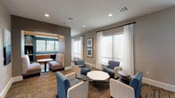 This single-floor business center is thoughtfully designed to accommodate both collaborative and focused work environments. The space is organized into distinct zones, beginning with an open lounge area furnished with a mix of contemporary blue upholstered chairs, neutral accent seating, and low-profile tables, including an eye-catching octagonal marble-top centerpiece. Patterned carpet in warm earth tones anchors the seating arrangements and adds visual texture throughout.

A semi-private alcove tucked into one corner offers an intimate setting with built-in shelving, accent lighting in a teal-green tone, and a small bistro-style table—ideal for focused individual work or quiet conversation. The main lounge transitions seamlessly into an adjacent area featuring leather lounge chairs and warm wood accent tables, lending a more relaxed, editorial feel.

Floor-to-ceiling black-framed glass partitions define the boundary between the business center and a neighboring common area, maintaining visual connectivity while providing a sense of separation. Multiple large windows dressed with sheer white drapery allow natural light to fill the space, creating a bright and welcoming atmosphere. Curated framed artwork adorns the walls, contributing to the polished, professional ambiance. Recessed lighting throughout ensures consistent illumination at all hours.