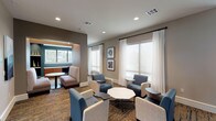 This single-floor business center is thoughtfully designed to accommodate both collaborative and focused work environments. The space is organized into distinct zones, beginning with an open lounge area furnished with a mix of contemporary blue upholstered chairs, neutral accent seating, and low-profile tables, including an eye-catching octagonal marble-top centerpiece. Patterned carpet in warm earth tones anchors the seating arrangements and adds visual texture throughout.

A semi-private alcove tucked into one corner offers an intimate setting with built-in shelving, accent lighting in a teal-green tone, and a small bistro-style table—ideal for focused individual work or quiet conversation. The main lounge transitions seamlessly into an adjacent area featuring leather lounge chairs and warm wood accent tables, lending a more relaxed, editorial feel.

Floor-to-ceiling black-framed glass partitions define the boundary between the business center and a neighboring common area, maintaining visual connectivity while providing a sense of separation. Multiple large windows dressed with sheer white drapery allow natural light to fill the space, creating a bright and welcoming atmosphere. Curated framed artwork adorns the walls, contributing to the polished, professional ambiance. Recessed lighting throughout ensures consistent illumination at all hours.