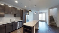 This two-story residence offers one bedroom, one and a half bathrooms, and a versatile den, delivering a well-appointed living experience with contemporary design throughout. The open-concept kitchen serves as a central hub, featuring dark espresso cabinetry that extends to the ceiling for generous storage, stainless steel appliances including a French door refrigerator and dishwasher, and a subway tile backsplash. A large granite-topped kitchen island provides additional prep space and casual seating, elegantly illuminated by a trio of pendant lights in varying modern silhouettes. Rich hardwood-style flooring flows seamlessly from the kitchen into the living area, where a ceiling fan and access to a private balcony through full-height glass doors invite natural light and outdoor views. Recessed lighting throughout enhances the bright, airy atmosphere of the main living spaces. The den adds a flexible area suitable for a home office or guest space, while the 1.5 bathrooms offer added convenience. Spanning two floors, this residence provides a sense of scale and separation rarely found in single-level layouts.