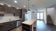 This two-story residence offers one bedroom, one and a half bathrooms, and a versatile den, delivering a well-appointed living experience with contemporary design throughout. The open-concept kitchen serves as a central hub, featuring dark espresso cabinetry that extends to the ceiling for generous storage, stainless steel appliances including a French door refrigerator and dishwasher, and a subway tile backsplash. A large granite-topped kitchen island provides additional prep space and casual seating, elegantly illuminated by a trio of pendant lights in varying modern silhouettes. Rich hardwood-style flooring flows seamlessly from the kitchen into the living area, where a ceiling fan and access to a private balcony through full-height glass doors invite natural light and outdoor views. Recessed lighting throughout enhances the bright, airy atmosphere of the main living spaces. The den adds a flexible area suitable for a home office or guest space, while the 1.5 bathrooms offer added convenience. Spanning two floors, this residence provides a sense of scale and separation rarely found in single-level layouts.