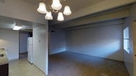 This single-floor space offers a straightforward and functional layout designed to make the most of its available square footage. The main living area is finished with wall-to-wall carpeting in a neutral tone, providing a comfortable and quiet underfoot experience. The walls are painted in a soft blue-gray hue, lending the space a calm and understated aesthetic throughout.

A defining feature of the living area is the multi-bulb chandelier with frosted bell-shaped glass shades and a brass-toned fixture, which serves as a central focal point and provides ample ambient lighting overhead. A structural beam runs along the ceiling, adding a subtle architectural detail to the room.

Adjacent to the living area, a transition to tile flooring marks the entrance to the kitchen zone, where a white refrigerator is visible, indicating a practical and accessible kitchen space. A light switch panel is conveniently positioned along the connecting wall between the two areas.

The space also features a visible floor vent for climate control, and the textured ceiling adds a classic residential finish. Overall, the unit presents a clean, move-in ready environment with a cohesive color palette and functional design suited for everyday living.