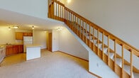 This two-story space is defined by its open, airy design and warm, functional finishes throughout. The main level centers on a generously proportioned living area with vaulted ceilings that amplify the sense of height and volume. A prominent staircase with classic wood railings and carpeted treads serves as a visual anchor, leading to the upper level while allowing sightlines to remain open between floors. A sliding glass door floods the living area with natural light and provides access to the outdoors. The kitchen is well-equipped with warm oak cabinetry, white appliances including a range, dishwasher, and refrigerator, and a peninsula that offers additional prep and seating space. Light tile flooring completes the kitchen's practical, clean aesthetic. Upstairs, the loft-style space benefits from a skylight and ceiling fan, creating a bright and comfortable retreat with an open railing overlooking the main level below. A carpeted bedroom on this floor includes a wide closet with double doors and a ceiling fan. The full bathroom features an oak vanity with ample storage, a combined tub and shower, and tile flooring, rounding out this well-organized, multi-level space.