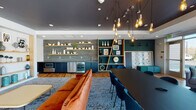 Located at 107 S River Rd in Bedford, New Hampshire, this single-floor communal space combines contemporary design with functional amenities. The open layout is anchored by warm hardwood flooring throughout, complemented by a bold color palette of teal, charcoal, and natural wood tones that give the space a vibrant yet cohesive feel.

The main lounge area features a striking built-in fireplace framed by open shelving with curated décor, alongside a seating arrangement centered on a plush orange sofa and a patterned area rug. A pendant lighting installation with exposed Edison bulbs adds an artful, industrial touch overhead.

The wet bar area is outfitted with a stainless steel beverage cooler, dark lower cabinetry, a sleek countertop, and a gray tile backsplash accented by floating natural wood shelves — ideal for hosting and entertaining. Adjacent to this is a large dining and gathering table surrounded by upholstered chairs, perfect for group collaboration or social events.

A window bar along one wall offers a long live-edge communal table lined with swivel barstools, bathed in natural light streaming through a row of floor-to-ceiling windows with views of the surrounding exterior. Near the entrance, a welcoming lounge nook with additional seating and a wall-mounted TV provides a comfortable retreat. Glass-paneled doors and large windows ensure the space remains bright and connected to the outdoors throughout.