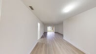 This single-floor space is designed with a clean, minimalist aesthetic throughout. The layout is open and expansive, with the main living areas finished in light-toned hardwood-style flooring that runs continuously, enhancing the sense of space. Crisp white walls and white baseboards maintain a bright, uniform appearance across all rooms.

The main living area is generously proportioned, with a sliding glass door providing direct access to an outdoor green space, allowing natural light to fill the interior. A second living or flex area connects seamlessly, offering views of a tranquil waterfront through a large window. Both spaces benefit from ceiling-mounted light fixtures and HVAC vents integrated overhead.

The bedrooms continue the light flooring theme. One bedroom includes mirrored sliding closet doors with interior rod and shelf storage. Another room features a built-in white cabinetry unit beneath a large window overlooking tropical landscaping and a palm-lined outdoor setting.

The bathroom is finished in large-format white tile flooring, featuring a single-sink vanity with a white countertop, white cabinetry with brushed hardware, a wall-mounted mirror, and standard fixtures. The overall ambiance throughout the space is clean, neutral, and move-in ready.