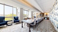 Located at 360 State St in New Haven, Connecticut, this thoughtfully designed single-floor space offers a refined and versatile environment suited for meetings, events, or collaborative gatherings. The centerpiece of the room is a striking long marble-top conference table flanked by plaid upholstered chairs and accented by elegant gold mushroom-cap table lamps, creating a sophisticated yet welcoming atmosphere. A gallery wall featuring an extensive curated collection of black-and-white photographs in gold frames lines one side of the room, adding a strong sense of place and visual interest. The entry corridor is distinguished by a series of artistically shaped gold-framed mirrors mounted against a cool blue-gray accent wall, lending a boutique-hotel aesthetic to the arrival experience. Floor-to-ceiling windows along the perimeter flood the space with natural light and offer open views of the surrounding New Haven skyline and greenery. A pair of deep navy wingback chairs positioned near the windows provides an inviting lounge nook. A built-in wooden bookshelf with display lighting anchors one end of the room, adding warmth and functionality. Neutral-toned patterned carpeting and crisp white ceilings with exposed beams complete the polished, contemporary design throughout.