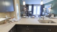 This single-floor residence showcases a thoughtfully designed open-concept layout that seamlessly connects the kitchen and living areas, creating a spacious and inviting atmosphere. The kitchen is a standout feature, boasting dark espresso cabinetry paired with speckled stone countertops that extend into a generous peninsula — ideal for casual dining or entertaining. A stainless steel sink and contemporary pendant lighting add polished, functional accents to the space. The kitchen opens directly into the living area, which is bathed in natural light streaming through large windows and elegant French doors dressed with dark curtain panels. The living room is warm and welcoming, with teal-accented walls providing a rich backdrop to the carefully curated furnishings, including plush seating and a rustic open shelving unit. The French doors hint at outdoor access, enhancing the airy, connected feel of the interior. Throughout the space, the interplay of dark and light tones — from the cabinetry to the countertops and wall colors — creates a balanced, modern aesthetic. The overall ambiance is both sophisticated and comfortable, making it a refined yet livable space suited for everyday living and hosting alike.