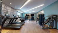 This fitness center is thoughtfully designed to offer a comprehensive workout experience within a polished, modern environment. The entry corridor sets an inviting tone with its mix of natural wood paneling, marble-style tile flooring, and a built-in upholstered bench framed in warm wood. A striking piece of abstract art mounted against a wood-paneled wall serves as a focal point, complemented by layered stone accent panels and recessed lighting overhead.

The main fitness floor features warm hardwood flooring and is outfitted with an extensive array of equipment, including multiple treadmills, stationary bikes, ellipticals, and a cable strength-training machine. A full dumbbell rack with rubber flooring provides a dedicated free weights area, while large framed mirrors visually expand the space and support proper form during training. Motivational athletic imagery is featured as a wall graphic, enhancing the energetic atmosphere.

A separate glass-enclosed studio area, accessible via sliding glass doors with a sleek wood-trimmed frame, offers a flexible space for stretching, yoga, or functional training, complete with a stability ball and open floor space. Large windows allow natural light to filter in throughout the facility, providing views to the surrounding streetscape. Linear LED lighting installed along the ceiling delivers bright, even illumination throughout the entire space.