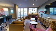 This thoughtfully designed lounge space offers a welcoming and sophisticated atmosphere through a carefully curated blend of furnishings and architectural details. The single-floor layout centers around a comfortable seating arrangement, anchored by a large neutral sofa adorned with patterned throw pillows in earthy gold and brown tones, complemented by richly upholstered burgundy accent chairs. A woven area rug grounds the seating area, adding warmth and texture to the hardwood flooring beneath.

Multiple round white-topped side tables are distributed throughout the space, providing functional surfaces while maintaining an open and uncluttered feel. Natural light floods the interior through a series of large windows and glass-paneled doors, offering views of lush outdoor greenery and creating a bright, airy ambiance. Recessed ceiling lighting supplements the natural illumination, ensuring consistent brightness throughout.

A decorative console table lines one wall, styled with pendant lighting above, fresh orchid arrangements, and ornamental accessories. A sculptural textured cabinet adds an artistic focal point near the entryway. The overall palette of warm yellows, grays, and deep reds creates an inviting and polished environment well-suited for relaxed social gatherings.
