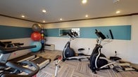 This fitness center is a thoughtfully designed, single-floor facility that provides a comprehensive workout environment. The interior is finished with a distinctive two-tone wall scheme — crisp white upper walls paired with a bold teal accent band — framed by dark wood crown molding that adds a polished touch throughout the space. Recessed lighting illuminates the room evenly, and large glass entry doors and windows allow natural light to flow in, offering views of the surrounding outdoor area.

The facility is well-stocked with a variety of cardio equipment, including multiple Spirit-brand treadmills, two elliptical cross-trainers, and a stationary spin bike, catering to a range of cardiovascular workout preferences. A dedicated free weights section features a multi-tier dumbbell rack, while a colorful stability ball rack holds an assortment of exercise and medicine balls. Additional equipment such as resistance bands and a weight bench further expand the training options available.

The flooring consists of durable, patterned carpet tiles that provide comfort underfoot during workouts. A wall-mounted mirror adds to the sense of space and supports form-checking during exercise. The overall layout is organized and functional, making efficient use of the available floor area to accommodate both cardio and strength training needs in a clean, welcoming environment.