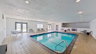This indoor pool facility is designed with functionality and comfort in mind, offering a well-maintained aquatic space suitable for year-round use. The centerpiece of the facility is a generously sized rectangular swimming pool with vivid turquoise water, framed by a classic dark tile border and a wide tiled surround that provides safe and spacious poolside access. Stainless steel entry railings make pool access straightforward and accessible.

The interior walls are finished in clean white and light grey tones, contributing to a bright, open atmosphere enhanced by recessed ceiling lighting. Multiple windows along the perimeter allow natural light to filter in, adding to the airy feel of the space. The flooring throughout the surrounding deck area consists of durable square tiles in neutral tones.

Seating is provided along the poolside in the form of contemporary metal chairs arranged in rows, along with a small table and chair set for casual use. A raised observation or utility area is visible along one wall, adding architectural interest to the space. Multiple entry and exit points, including double-door access, ensure convenient circulation throughout the facility. Overall, the space presents a clean, practical, and welcoming environment.