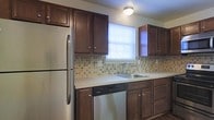 This single-floor residence offers a thoughtfully designed layout that balances functionality and comfort. The kitchen serves as a central highlight of the space, showcasing rich dark-stained cabinetry that extends from floor to ceiling, providing generous storage throughout. Light-toned countertops offer a clean contrast against the darker woodwork, while a decorative mosaic tile backsplash in warm earth tones adds visual texture and character to the space. Stainless steel appliances, including a full-size refrigerator and dishwasher, complement the kitchen's practical design. A stainless steel sink is positioned beneath a window fitted with white blinds, allowing natural light to filter into the kitchen and brighten the workspace. Overhead, a flush-mount ceiling light fixture ensures even illumination throughout the room. The overall ambiance of the space is warm and inviting, with the combination of dark cabinetry, neutral countertops, and earthy backsplash tones creating a cohesive and timeless aesthetic. The single-floor layout contributes to an easy, accessible flow throughout the home, making it a practical and comfortable living environment.