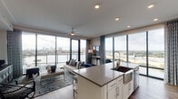 This elegant high-rise residence offers a seamlessly designed single-floor layout that maximizes its stunning waterfront and city skyline vistas. Floor-to-ceiling glass windows and sliding doors span the living areas, bathing the interiors in natural light and framing breathtaking panoramic views of the water and surrounding cityscape.

The open-concept living and dining area is anchored by a generous seating arrangement and connects effortlessly to the gourmet kitchen, which features crisp white cabinetry, a marble-style backsplash, stainless steel appliances including a French door refrigerator and wine cooler, and a large center island with quartz countertops and bar seating. Warm hardwood flooring flows throughout the main living spaces.

The primary bedroom offers direct access to the balcony through floor-to-ceiling glass doors, along with plush carpeting, ample storage via a multi-drawer dresser, and curated artwork. The en-suite bathroom impresses with a spacious walk-in glass shower enclosed in white tile, a wood bench, and a long vanity with dual under-mount sinks and generous cabinetry. A second bedroom provides the same refined comfort with carpet flooring, a ceiling fan, and waterfront views. The residence also features a well-appointed full bathroom and private balcony overlooking the water.