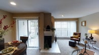 This single-floor residence offers a welcoming and well-organized layout centered around a standout fireplace with a tiled hearth and white mantle, serving as the focal point of the main living area. The living room is generously proportioned, with plush carpeting underfoot and large windows dressed in vertical blinds that allow abundant natural light to filter through. A wide sliding glass door opens to an outdoor patio area, where outdoor seating is visible, extending the sense of space and providing easy indoor-outdoor access. The warm, neutral-toned walls complement the open feel of the room, while recessed lighting adds a clean, functional touch to the ceiling. A dining area is situated adjacent to the living space, partially visible with dark wood furnishings that contrast the lighter tones throughout. The fireplace is flanked by decorative greenery and personal accents atop the mantle, lending a homey character to the space. The combination of tile at the hearth and carpet in the living area creates a natural visual separation of zones. Overall, the space presents a comfortable, functional, and light-filled single-floor home with a cohesive design aesthetic.