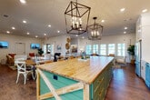 This single-story space is designed with a modern farmhouse aesthetic that blends functionality and style across its open, interconnected layout. The centerpiece of the space is a generously sized kitchen featuring a standout butcher block island with teal barn-style paneling and cross-brace detailing, suspended above by striking black geometric cage pendant lights. White shaker cabinetry, subway tile backsplash, open wood shelving, and stainless steel appliances complete the kitchen's polished yet rustic character.

The kitchen flows seamlessly into a spacious open living and dining area finished with rich hardwood flooring throughout. Multiple seating arrangements, round dining tables with cross-back chairs, and lounge furniture create flexible gathering spaces bathed in natural light from rows of large windows along the perimeter walls.

A dedicated lounge nook features a bold green shiplap accent wall with built-in cabinetry and open shelving, serving as a beverage station and casual retreat. An adjacent sitting area includes wall-mounted televisions and comfortable seating, ideal for relaxed entertainment.

The fitness room is well-appointed with cardio equipment including a treadmill, elliptical, and stationary bikes, complemented by wall-to-wall mirrors, recessed lighting, and multiple windows providing natural light and outdoor views.