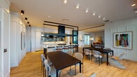 The Social Lounge is a thoughtfully designed communal space that seamlessly integrates multiple functional zones within a single open-plan layout. The interior is characterized by warm hardwood flooring, white walls, and a carefully curated palette of black architectural accents that define each area without enclosing it.

At the heart of the space is a fully equipped demonstration kitchen, outfitted with a gas cooktop island, extensive white cabinetry, and a bold black overhead hood structure that serves as a striking architectural focal point. Mirrored surfaces along the kitchen wall amplify the sense of space and light. A bar-height counter with seating provides an ideal vantage point for cooking demonstrations or casual dining.

The dining area features multiple dark-topped tables with a variety of contemporary chair styles, accommodating both small gatherings and larger groups. A wall-mounted display screen adds a media component to this zone.

Adjacent to the kitchen, the lounge area offers plush gray sofas, rust-orange accent chairs, statement floor lamps, and a media console with a large flat-screen television, all anchored by area rugs for an intimate atmosphere.

Floor-to-ceiling glass doors open directly onto an outdoor terrace, flooding the interior with natural light and providing views of the surrounding cityscape and greenery.
