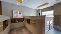 This well-appointed mailroom and lobby space serves as a central hub for residents, combining practical functionality with a clean, modern aesthetic. The area is defined by its large-format tile flooring, which transitions between warm taupe and cool grey tones across different zones of the space, lending a polished, contemporary feel underfoot.

A prominent central counter with a stone-topped surface anchors the room, providing a convenient surface for sorting and handling deliveries. Multiple banks of individual mailboxes line the walls and the central island, finished in both bronze and champagne tones, offering organized mail access for a large number of residents. A dedicated parcel locker unit provides secure storage for package deliveries.

A framed community bulletin board is mounted on the wall, keeping residents informed of local notices and events. The space is illuminated by a striking arrangement of pendant lights suspended from a distinctive gold-accented ceiling feature, adding a warm, decorative touch to the otherwise neutral palette of taupe and white walls.

Expansive floor-to-ceiling windows along one side of the lobby flood the area with natural light and frame views of the surrounding greenery and landscaping, creating a bright and welcoming atmosphere. The overall design balances utility and style, making this an efficient and pleasant space for daily resident use.