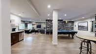 This single-floor amenity space is a thoughtfully designed common area that blends comfort, entertainment, and social functionality. The open layout is anchored by striking stacked-stone accent walls in a dark grey palette, lending an upscale, lodge-inspired character to the room. Durable wood-look flooring extends across the main entertainment zone, while patterned carpet defines the lounging and dining areas.

At the heart of the space is a classic billiards table with a vibrant blue felt surface, complemented by high-top round tables and industrial-style stools — ideal for casual gatherings. A dedicated lounge area features a set of tufted leather Chesterfield sofas arranged around a rustic wood coffee table, creating an inviting setting for conversation and relaxation. Recessed lighting throughout ensures the space is well-lit, and a wall-mounted mini-split unit provides climate control.

A compact kitchenette with white shaker cabinetry, a subway tile backsplash, granite countertops, and a stainless steel dishwasher adds practical convenience. Adjacent to the kitchenette, a casual dining nook with a round table and cross-back chairs provides an informal setting for meals or activities. Curated artwork, including bold equestrian paintings and framed prints, is displayed throughout, contributing to the space's warm and characterful ambiance.