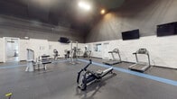 This fitness facility is designed to support a comprehensive workout experience across a single, thoughtfully organized floor. The main exercise area is expansive, featuring soaring ceilings with high-set windows that allow natural light to filter in, complemented by overhead lighting and a large ceiling fan that promotes air circulation throughout the space. The floors are covered in durable rubber matting with blue foam flooring sections designated for stretching and functional training, including an agility ladder for speed and coordination drills. The equipment selection is diverse, including Matrix-brand rowing machines, multiple treadmills, ellipticals, stationary bikes, a cable and pulley strength machine, a power tower, and a chest press machine. Wall-mounted flat-screen televisions provide entertainment throughout the workout floor. A vibrant accent wall featuring multicolored horizontal planks in blue, teal, and neutral tones adds a striking visual element to one section of the room, while other walls are finished with white shiplap-style paneling against a dark upper wall and ceiling. A separate entry room features warm hardwood flooring, white wainscoting, and decorative horizontal striping in green and gray, providing a welcoming transition into the facility. Full-length mirrors along one wall enhance the sense of openness and support form-checking during exercise.