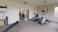 This single-floor fitness room is a comprehensively equipped exercise space designed to support a wide range of workout routines. The room is finished with neutral beige walls and wall-to-wall carpet, with rubber mats placed beneath cardio equipment for added protection and stability.

A row of large windows along one wall floods the space with natural light and offers views of surrounding greenery, creating a bright and inviting atmosphere. Cardio equipment includes a treadmill, a recumbent stationary bike, and an elliptical trainer. Strength training is supported by a multi-station cable machine mounted along one wall, a dumbbell rack with a tiered set of free weights, and an adjustable workout bench. Additional fitness accessories include a stability ball, a foam roller, a yoga mat, and a wall-mounted flat-screen television for entertainment during workouts.

The room also features a coat rack near the entrance, a wall clock, and a closet for storage. An adjacent space, visible through a glass-paneled door, appears to serve as a changing or utility area, adding functional convenience to the overall layout. The room is well-ventilated with ceiling vents and maintains a clean, organized environment throughout.