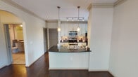 Located at 5201 Collin-McKinney Pkwy in McKinney, TX, this single-floor residence offers a thoughtfully designed layout with stylish finishes throughout. The kitchen is a standout feature, showcasing rich dark granite countertops, crisp white cabinetry extending to the ceiling, and a full suite of stainless steel appliances including a range, microwave, and refrigerator. A curved breakfast bar peninsula with pendant lighting separates the kitchen from the living area, creating an open and functional flow. Track lighting and a tiled backsplash add polished detail, while a window provides natural light and an outdoor view. The living areas feature warm, dark hardwood flooring that ties the open-concept space together seamlessly. Crown molding adds an elegant architectural touch throughout. The bedroom is generously sized with plush neutral carpeting, multiple windows dressed with blinds, and a ceiling fan with light fixture for year-round comfort. The bathroom is clean and functional, featuring a tub-shower combination with white tile surround, granite-topped vanity, large mirror, and tile flooring. Overall, the space presents a cohesive, modern aesthetic with quality finishes, making it a comfortable and attractive residence in the heart of McKinney.
