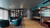 Located at 30 W First Ave in Columbus, Ohio's vibrant Short North neighborhood, this single-floor amenity space combines sophisticated design with functional gathering areas. Light hardwood flooring flows throughout, unifying the distinct zones within the open layout.

The lounge area anchors the space with tufted leather sofas, an upholstered ottoman, and curated wall art set against a textured dark accent wall. Sconce lighting and recessed ceiling fixtures create a warm, gallery-like ambiance, while large glass doors open directly to an outdoor patio, filling the room with natural light.

Adjacent to the lounge, a pair of plush teal swivel chairs surround a round side table, set against an open wood-and-metal shelving unit styled with decorative accents and table lamps—offering an inviting spot for casual conversation or quiet reading.

The café-style bar area is a standout feature, showcasing a rich walnut countertop trimmed with black herringbone tile, industrial-style pendant lighting, and a row of round wood-seated barstools. A farmhouse sink, open metal shelving, and a coffee station complete this well-appointed space.

A private dining nook with upholstered booth seating and a compact table adds an intimate dining option. The overall palette of teal, charcoal, and warm wood tones gives the space a cohesive, contemporary character.