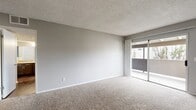 Located at 10522 Santa Gertrudes Ave in Whittier, CA, this one-bedroom, two-bathroom unit offers a well-organized single-floor layout designed to maximize functionality. The combined kitchen and living area serves as the heart of the unit, with the galley-style kitchen featuring rich dark cabinetry extending to the ceiling, light stone countertops, a stainless steel sink, and black stainless appliances including a gas range and refrigerator. Wood-style hard flooring runs throughout the kitchen, providing a clean, durable surface. The living room is finished with plush carpeting and neutral gray walls beneath a textured ceiling, creating a calm and inviting atmosphere. Sliding glass doors flood the space with natural light and open directly to an outdoor balcony, offering views of the surrounding greenery. The spacious primary bedroom also features carpeted floors, soft neutral tones, and its own sliding glass door access to a private outdoor area. A hallway connects the bedrooms and bathrooms, lined with full-length mirrored closet doors that enhance the sense of space. Both bathrooms are finished with dark vanity cabinetry complementing the overall design palette of the unit.