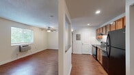 This efficient and thoughtfully laid-out 1-bedroom, 1-bathroom unit offers a comfortable living experience across a single floor. The combined kitchen and living area forms the heart of the unit, featuring rich hardwood-style flooring that extends throughout the main living spaces. The kitchen is equipped with warm wood cabinetry, granite-look countertops, and a full suite of black appliances including a range, dishwasher, and refrigerator. Recessed lighting illuminates the kitchen, while a partial dividing wall creates a natural separation between cooking and living areas. The living room is bright and open, benefiting from a large window that invites natural light and outdoor views, complemented by a ceiling fan overhead. A wall-mounted HVAC unit provides individual climate control. The bedroom continues the comfortable feel with plush carpeting, a ceiling fan, a window with outdoor views, and a wide sliding-door closet with wire shelving for ample storage. The bathroom is a standout feature, showcasing large-format marble-style tile surrounding a full soaking tub and shower, finished with sleek dark hardware. A dedicated closet space with wire shelving and carpet flooring adds further practical storage to the unit. Textured ceilings are present throughout.