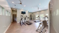 This dedicated fitness center offers a thoughtfully designed workout space that accommodates a comprehensive range of exercise equipment. The room features light sage-green walls, durable wood-look flooring, and a clean, open layout that maximizes usable floor space for movement and training.

Natural light fills the room through a series of windows, including a distinctive row of horizontally oriented clerestory windows along one wall that frame views of surrounding greenery. Two ceiling fans provide consistent airflow throughout the space, complementing the recessed lighting that ensures bright, even illumination at all hours.

The cardio zone is well-appointed with multiple treadmills, an elliptical trainer, and a stationary bike, while the strength training area includes a multi-function cable machine, a weight bench, and additional resistance equipment. A stability ball adds flexibility training options to the mix. A wall-mounted flat-screen television is positioned for easy viewing during workouts. Mirrors along one wall enhance the sense of openness and allow users to monitor their form. A fire extinguisher is also visibly mounted for safety. The single-level layout and dedicated entry door make the space easily accessible, creating a functional and inviting environment for fitness.