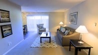 This single-floor space offers a comfortable and well-appointed layout designed for easy, everyday living. The living area serves as the heart of the space, featuring soft wall-to-wall carpeting and a neutral color palette that creates a calm, welcoming atmosphere. Large sliding glass doors fitted with vertical blinds allow generous amounts of natural light to fill the room, enhancing its open and airy feel. The living area provides ample space for seating and relaxation, with room for a sofa, accent chair, and a central coffee table arranged over a patterned area rug. Framed artwork on the walls adds a finishing touch to the space. Adjacent to the living area, a dining space is visible, accommodating a small table and chairs — ideal for casual meals. The overall design is clean and functional, with smooth walls, standard-height ceilings, and consistent flooring throughout. The neutral tones and soft furnishings contribute to a cozy yet understated ambiance, making this a practical and livable space suited to a variety of lifestyles.